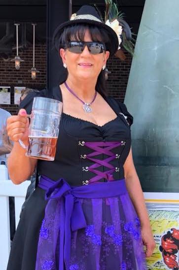 Woman wearing a black and purple dirndl holding a beer at an event. She is wear a bavarian hat and edelweiss necklace
