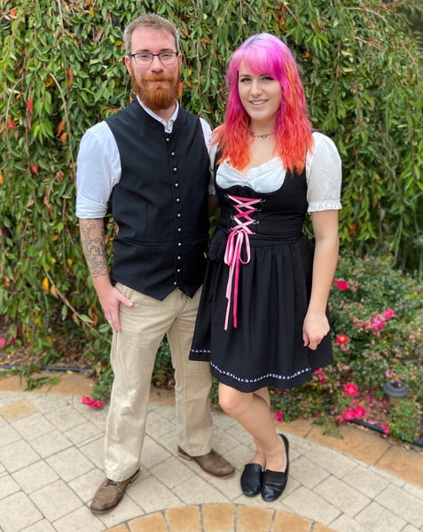 man in an authentic trachten vest standing next to a woman with pink hair wearing a white blouse and black traditional dirndl at an german festival