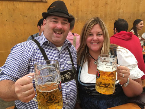 man wearing lederhosen sitting next to a woman wearing a plus size dirndl holdins liter beer steins at Oktoberfest