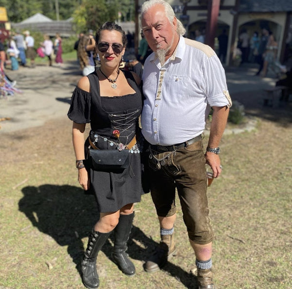 Woman wearing an all black Dirndl with tall leather boots, standing next to a man and lederhosen