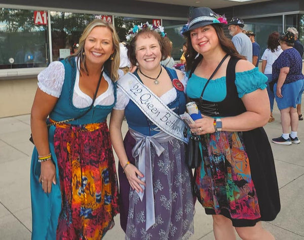 Group of women in traditional garments or German dirndl from the alpine regions for Oktoberfest. One woman is in a while blouse with bloue dirndl and colorful apron, one woman is in a white cotton blouse, blue dirndl bodice, with purple apron and gray embroidery, and gray ribbon lace up. The last woman is in a hat with an off the shoulder cotton apron, black german dirndl dress, black bodice and colorful apron.