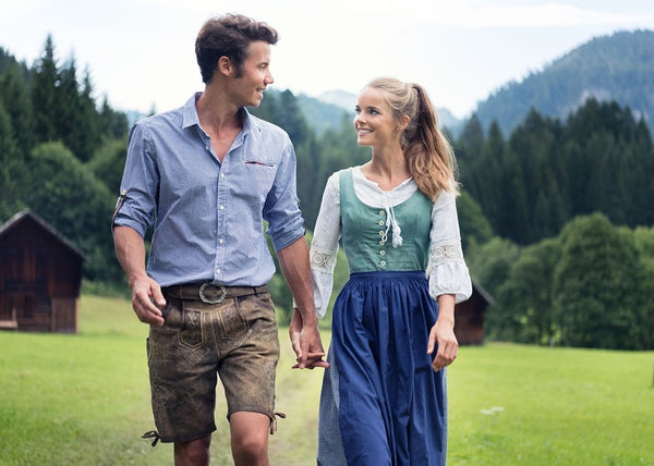 A man in lederhosen and a blue shirt, holding hands with a girl wearing a Dirndl with a long sleeve white blouse 