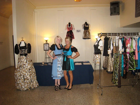 two women wearing German dirndl dresses, one is sky blue  and the other is a turquoise blue posing next to clothing racks of contemporary dirndls