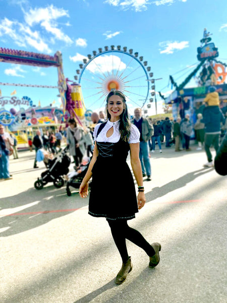 Woman with long brown hair, wearing a black Dirndl and white blouse in front of the rides at Oktoberfest