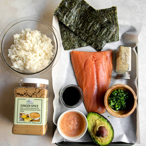 Ginger powder in clear tub. Salmon filet. half an avocado. green onion. seaweed. sesame seed. all on a white countertop.