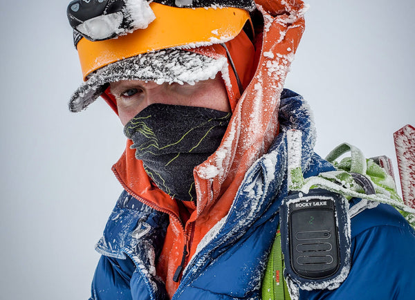 A mountaineer with a blue jacket, orange helmet, and Rocky Talkie squints through the rime ice covering him.