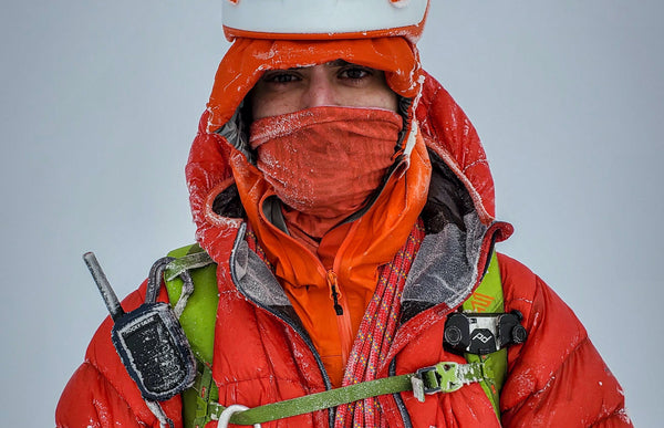 A mountaineer with a red jacket, green backpack, and Rocky Talkie is covered with frost and snow. 