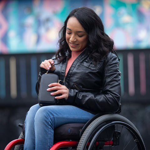 A young adult sitting in her wheelchair holding the FFORA essentials bag in black.