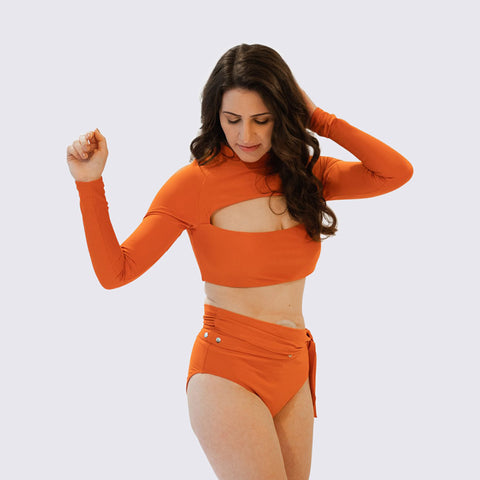 Woman standing centre frame in orange Lydia swim top & bottom by Miga Swimwear on a grey background.