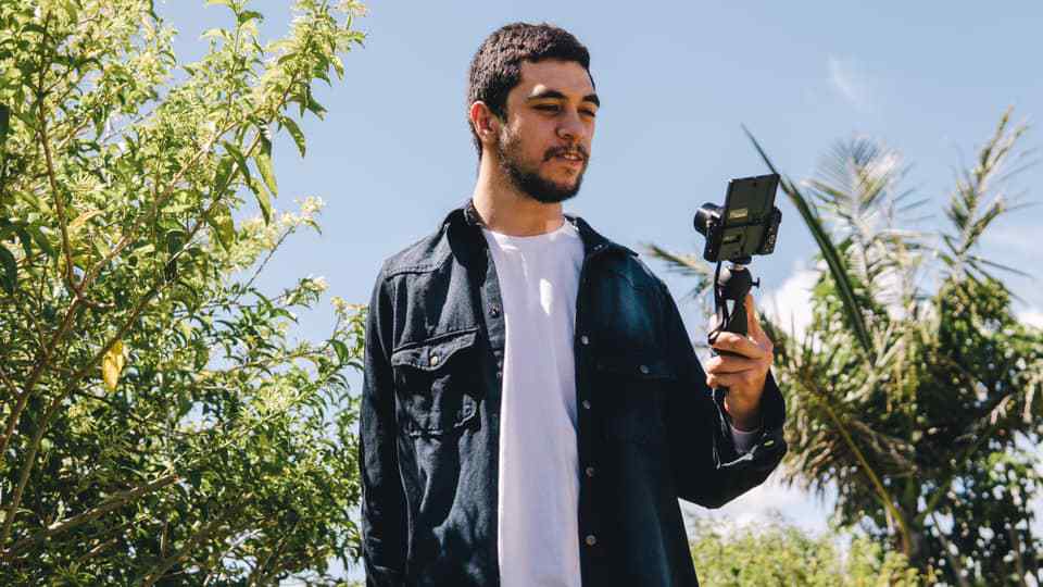 Will, a young NZ man is holding a selfie stick filming himself. He is in a garden. 