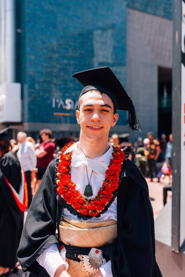 William, a young NZ man is wearing a graduation gown with traditional cultural attire.