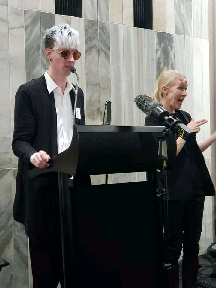 Ari, a young man is standing on a podium speaking into a microphone next to a sign language interpreter. He is wearing brown shades, white button up shirt and a black jacket. Ari has a mullet haircut, bleached silver at the top. 