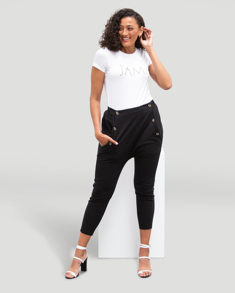 Alt text: Woman wearing High Waisted pant in black and white tee & leaning against a white podium & a grey background.