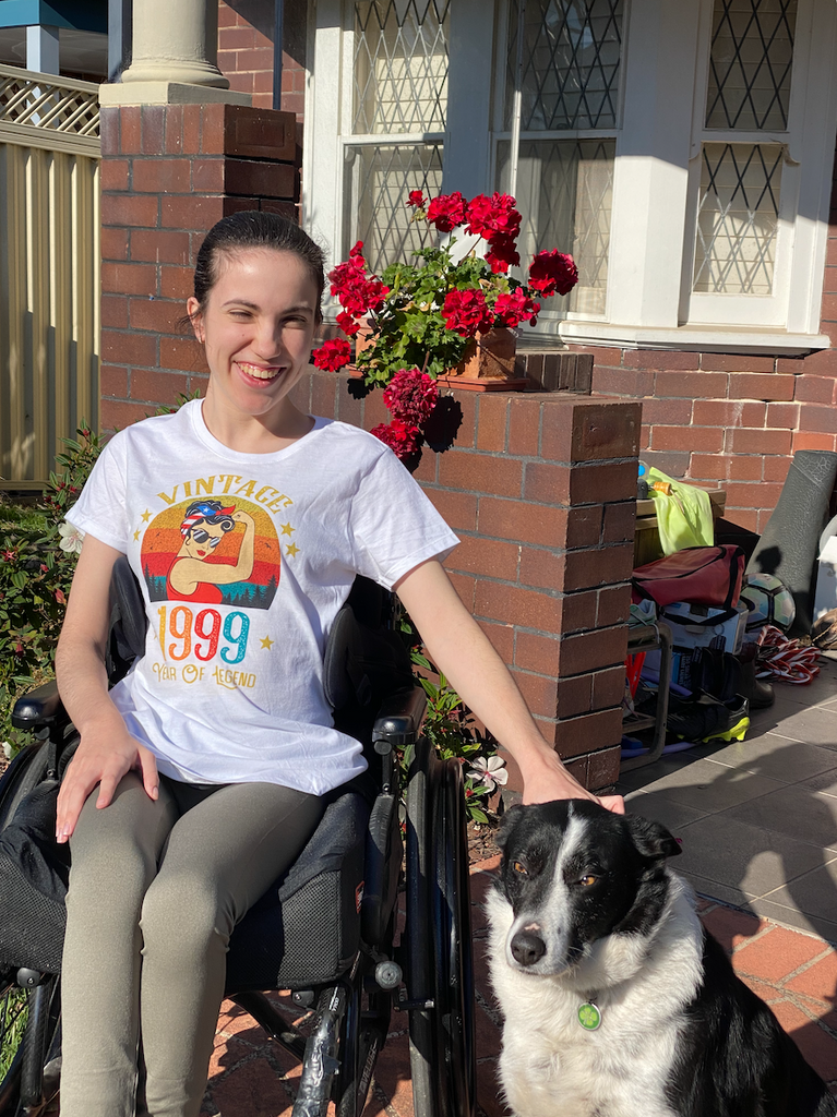 Hannah is wearing a graphic white tee saying "vintage 1999 year of legend" with a feminist theme. She is outside in a front garden patting a dog. She's using a wheelchair.