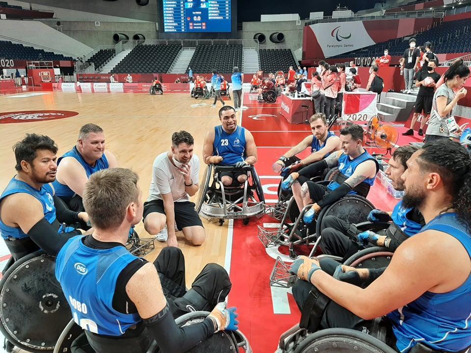 Barney and his team the Wheelblacks at the Tokyo 2020 Paralympic Games on-court gathering around in a circle. He is with seven teammates who are wearing blue jerseys and his coach is wearing a white tee and black shorts crouching on the ground. 