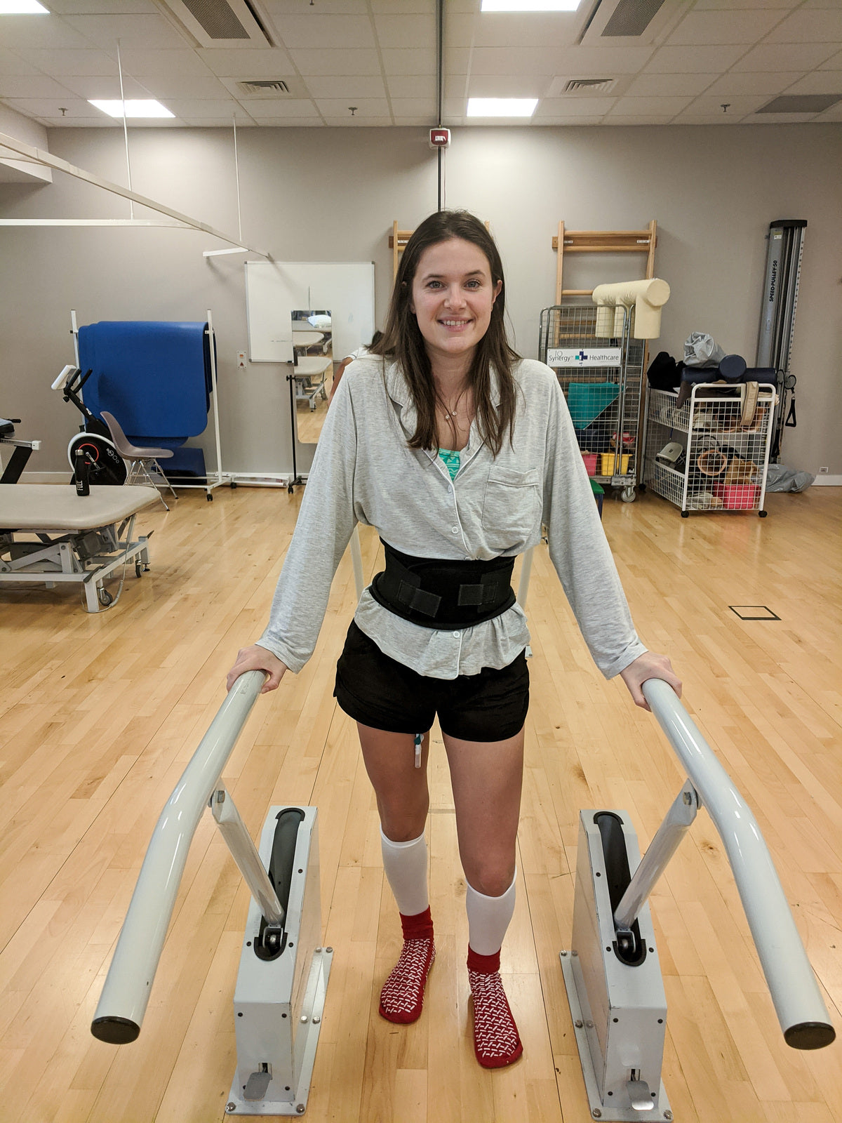 Alt text: A front shot of Sarah standing on her own, relearning to walk during a rehabilitation session & with a smile on her face.