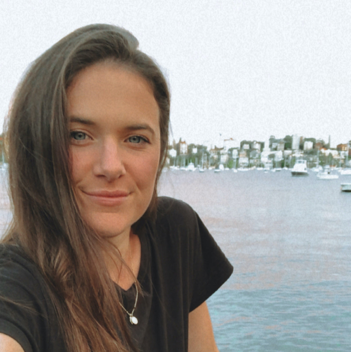 Alt text: A selfie of Sarah wearing a black tee with the ocean, boats and shoreline in the background over her left shoulder.