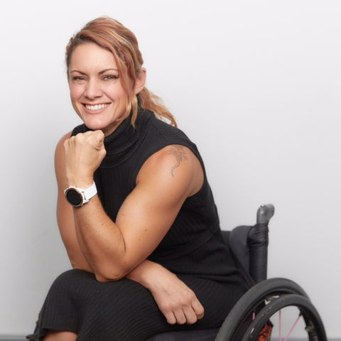 Natasha, a caucasian woman is smiling with her head resting on her hand, using a black wheelchair.