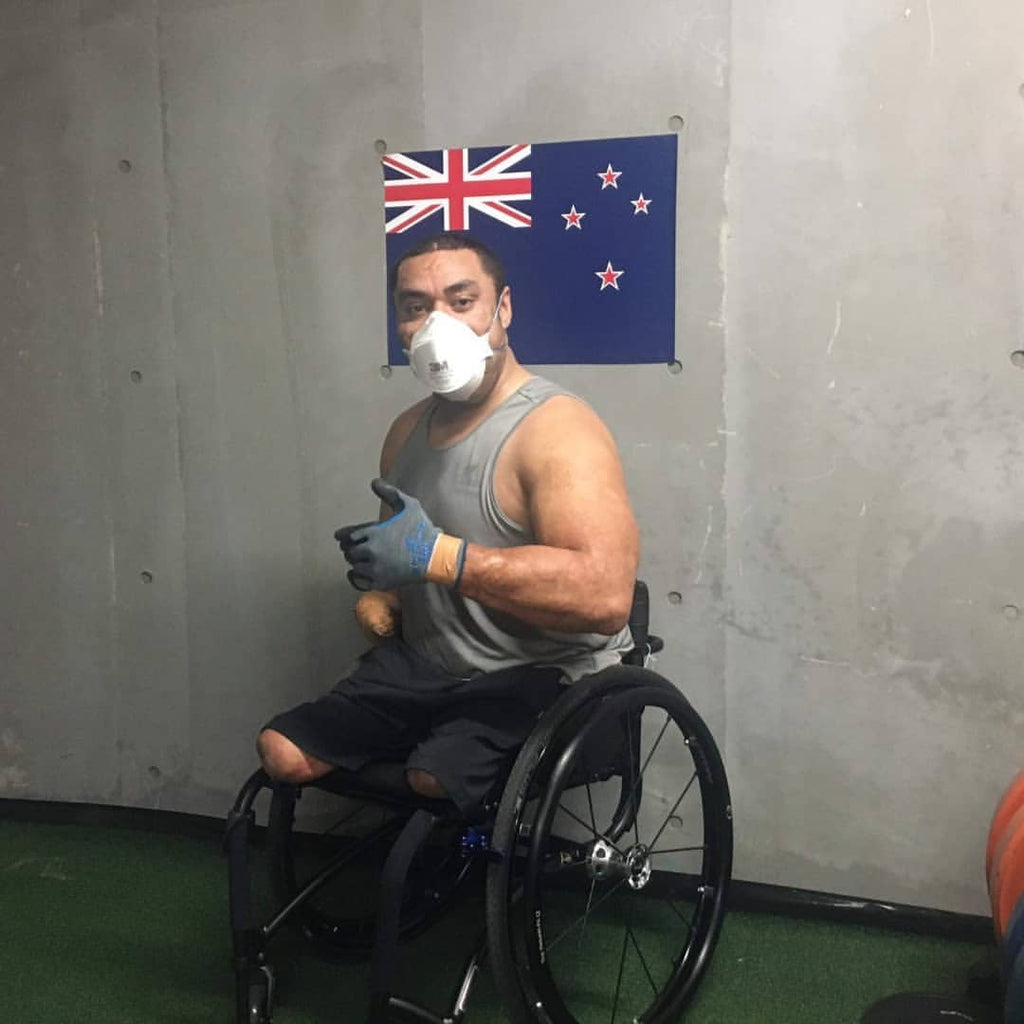 Barney, a young NZ man is wearing a grey tank and black shorts with his thumbs up wearing a fitted mask. He is using a wheelchair sitting in front of the NZ flag.