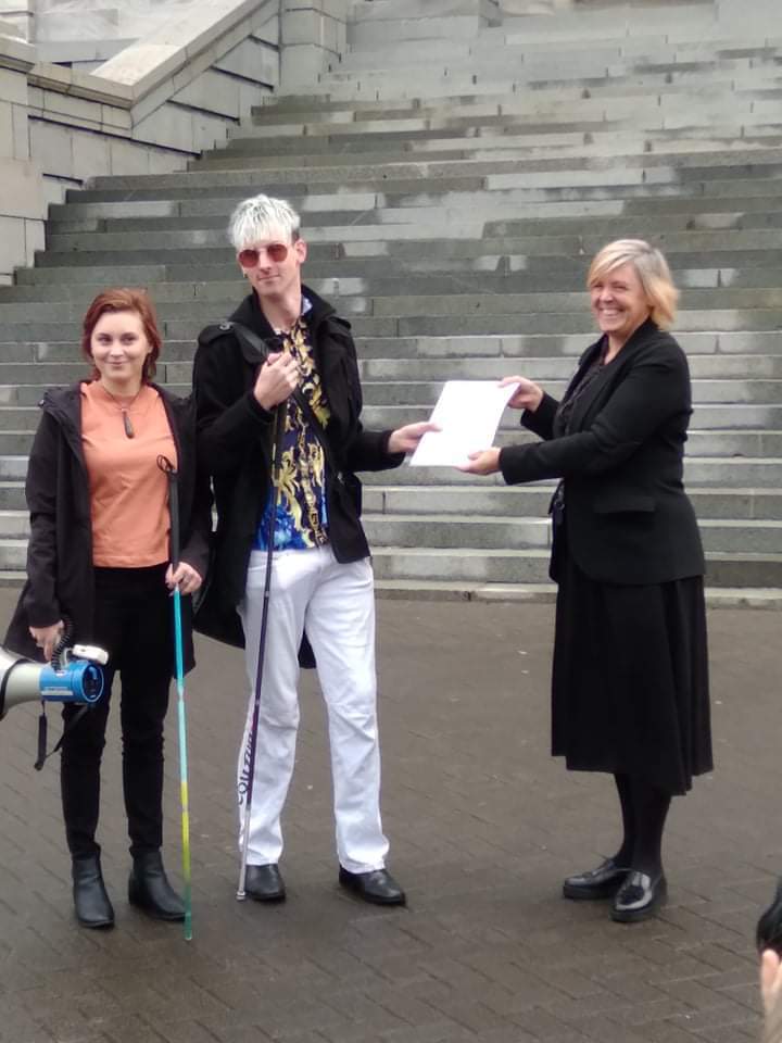 Ari and Karley are standing in front of concrete stairs posing for a photo. Ari is holding a document with a government official and Karley is on his right, holding a loudspeaker and her blue and yellow cane. Both are smiling for the camera.