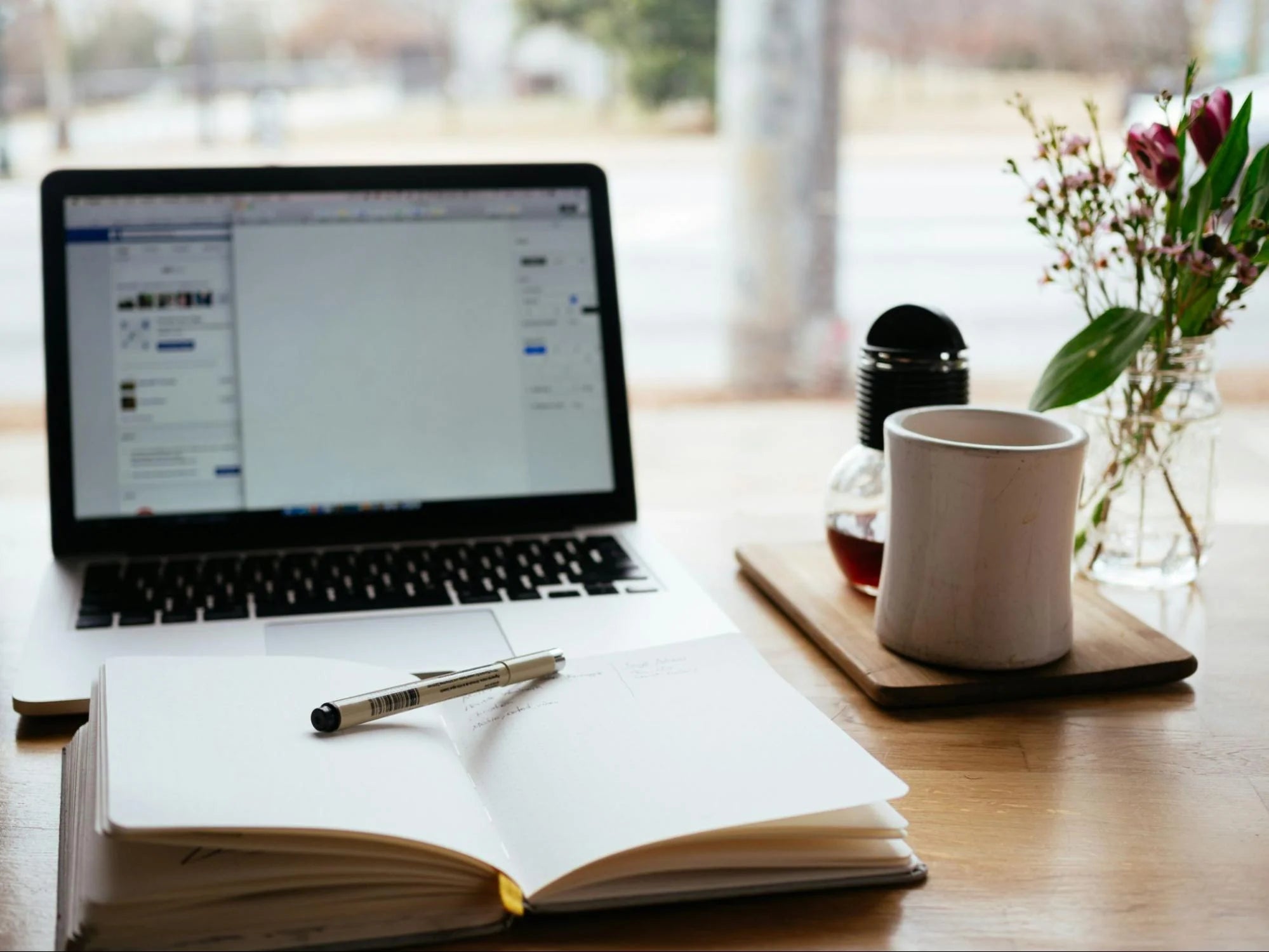 Un ordinateur ouvert sur Facebook, à côté d'une tasse de café, d'un carnet ouvert et de fleurs