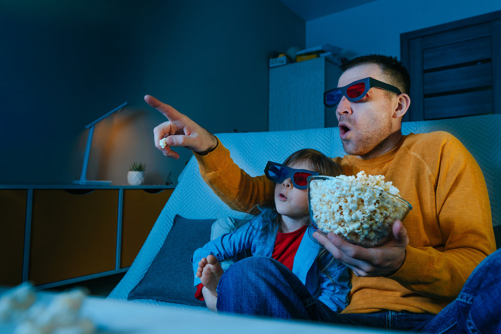 Father in yellow sweater and son in red and blue jacket watch movie. Father holds popcorn bowl and points at tv. Both are wearing 3D movie glasses.