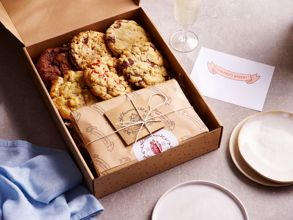 Variety Box Chummys Cookies wrapped in brown paper with small plates to the right and below. A blue napkin to the left.