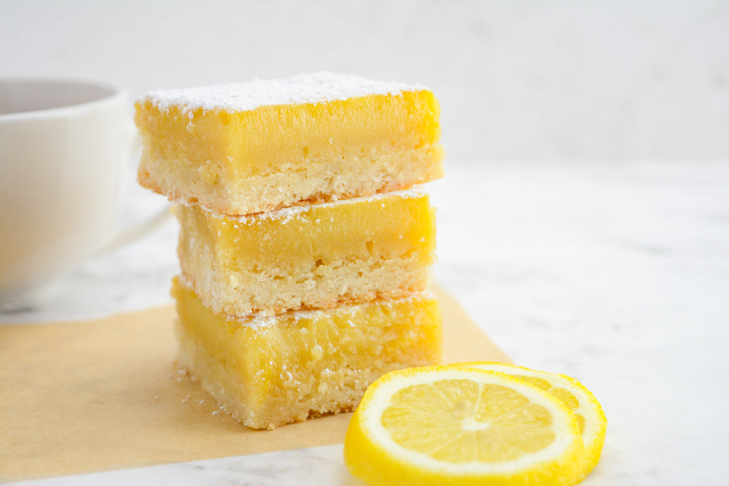 Three stacked lemon bars on brown parchment paper. In front is a lemon slice and behind is a tea cup.