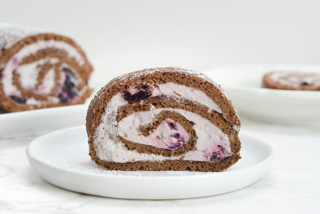 Chocolate rolled cake with pink cream spiral. Slice sis on plate in center with whole cake and another single serving behind.