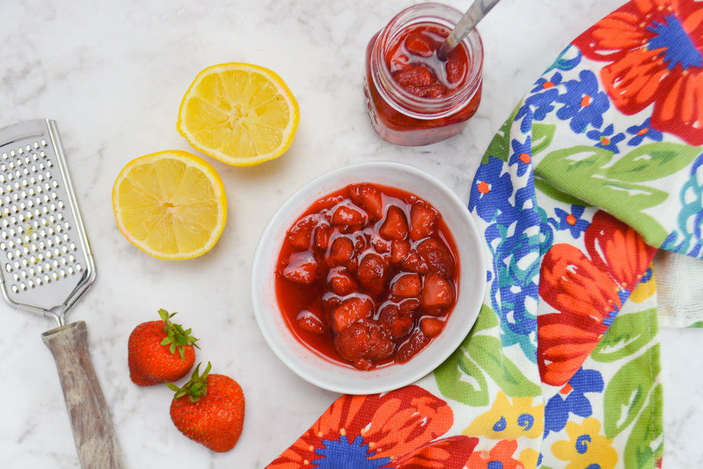 White bowl of strawberry compote is surrounded by lemons, strawberries and a grate to the left, a colorful blue and red napkin to the right
