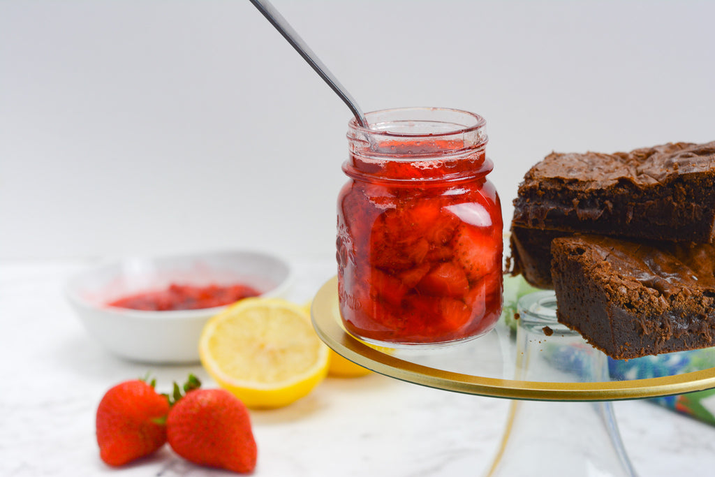 A jar of compote sits on a gold rimmed cake stand. To the left is a bowl, lemons and strawberries. A stack of brownies to the right