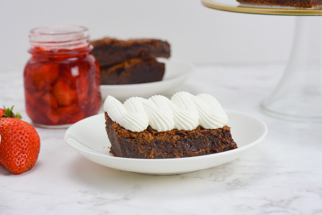 Brownie with whipped cream topping sits on a white plate with a jar of compote to the left