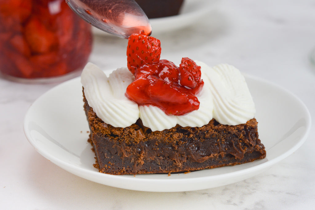 A brownie sits on a white plate with whipped cream. A spoonful of strawberry compote is being added