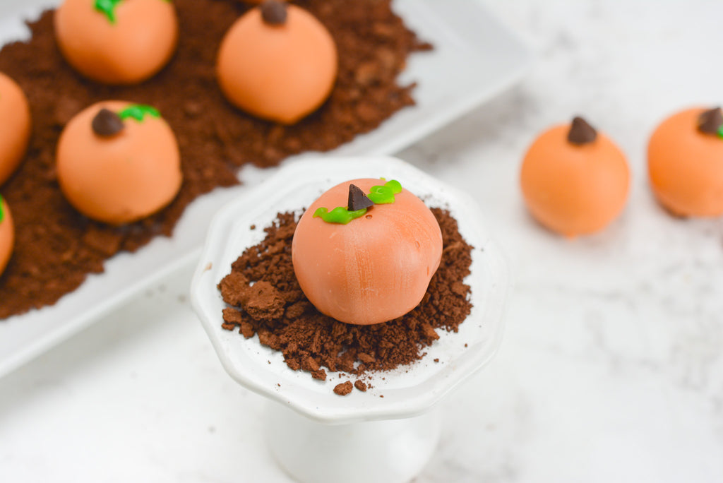 Pumpkin blondie truffles sit in graham cracker dirt to look like a pumpkin patch. A single pumpkin truffle sits towards the front on a cupcake stand.