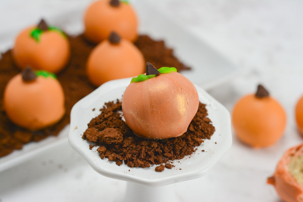 Pumpkin blondie truffles sit in graham cracker dirt to look like a pumpkin patch. A single pumpkin truffle sits towards the front on a cupcake stand brushed with glittering luster dust