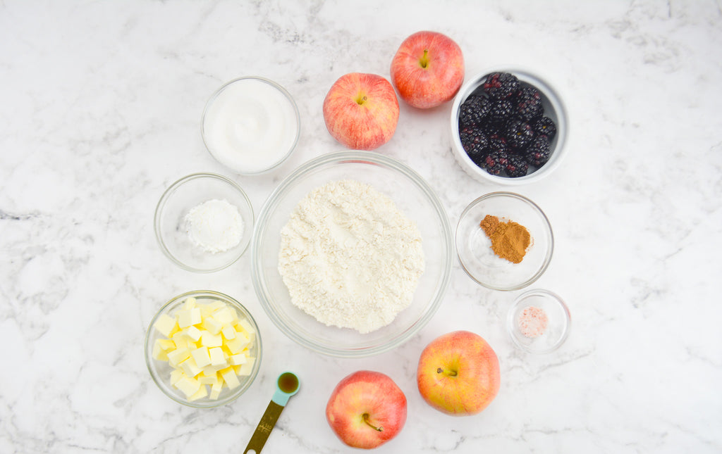 A bowl of flour is surrounded by bowls of the the ingredients for the crumble and fresh apples