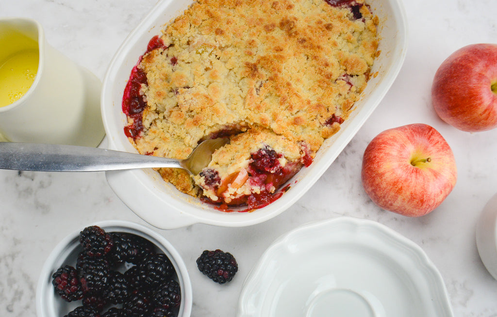 A baking dish of crumble with a spoon taking out a serving. A white plate sits towards the bottom and blackberries, fresh apples and custard surround
