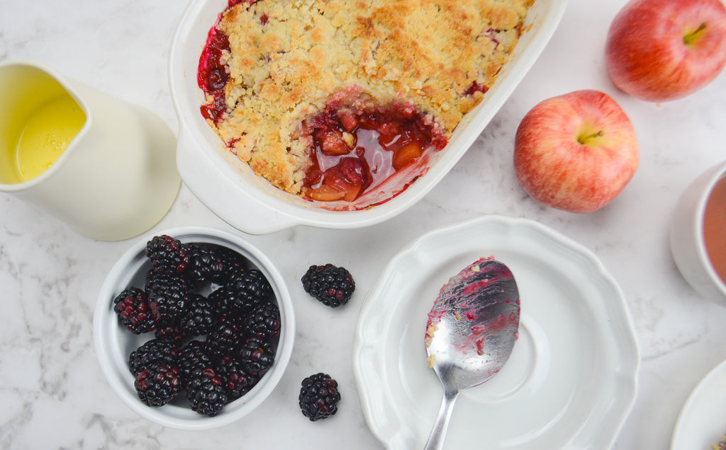 A full casserole dish has a spoonful taken out exposing the dark red filling. A spoon with filling sits underneath and a bowl of blackberries and custard to the side.