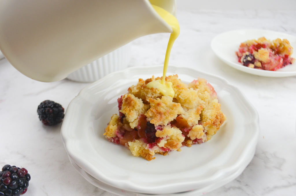 A small pitcher of custard is poured on top of a single serving of fruit crumble