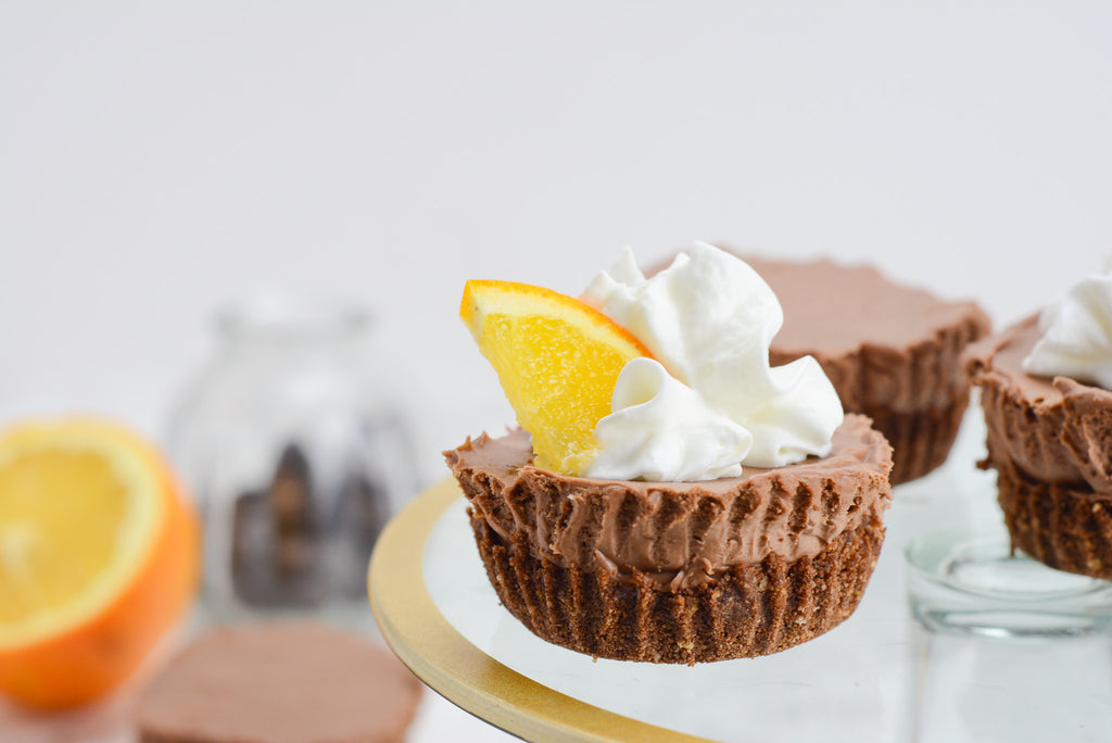 Mini chocolate cheesecake sits on a gold rimmed cake stand with whipped cream and orange slice as garnish. chocolate chips and an orange are in the background.