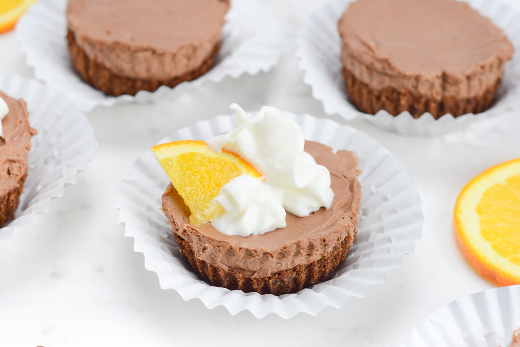 A mini cheesecake sits inside of a cupcake liner and is topped with whipped cream and an orange slice