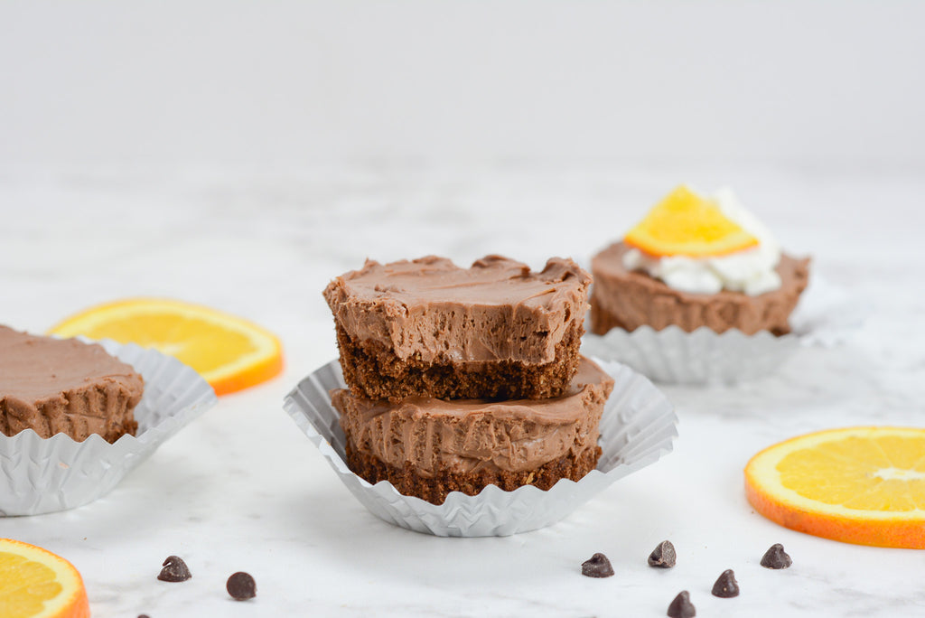 Two mini cheesecakes are stacked with a bite taken out of the top one. orange slices, chocolate chips and other cheese cakes fill the background