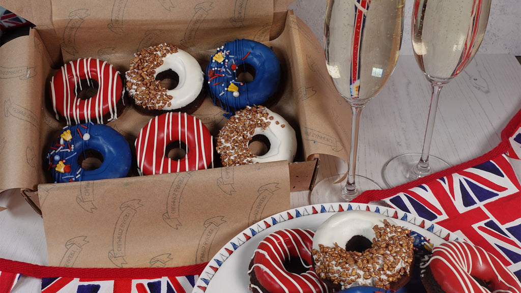 Brown box of red white and blue bronuts on a white table adorned with flags and two champagne flutes and a plate of stacked decorated bronuts