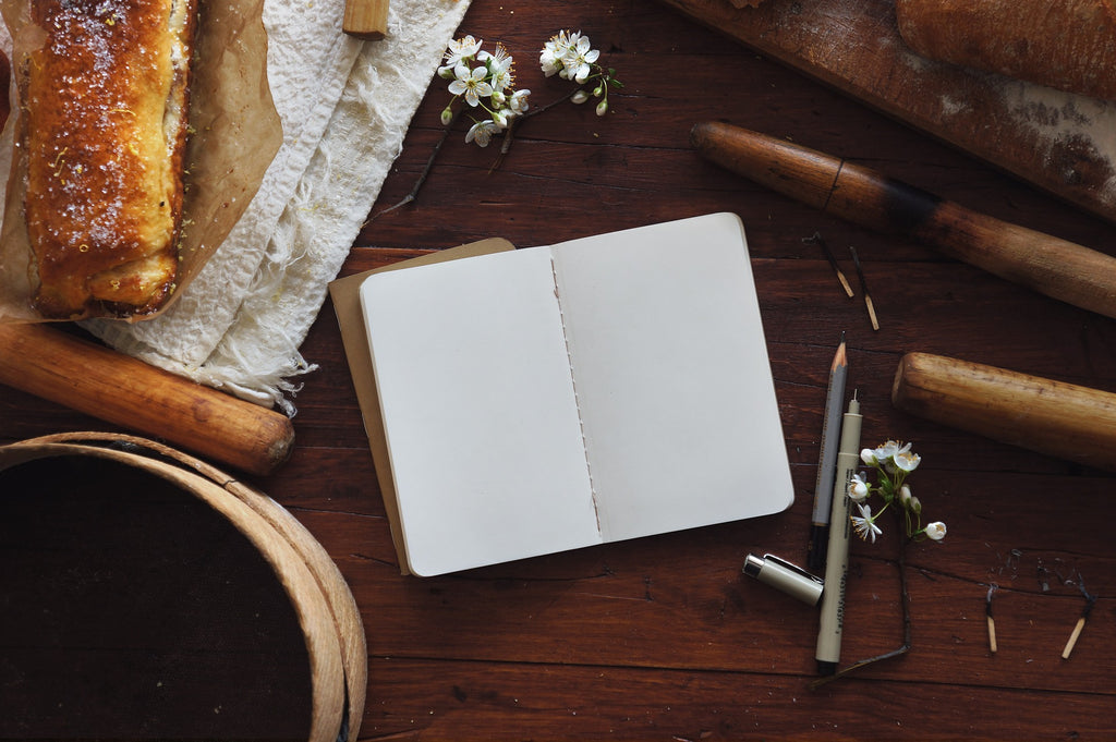 Open blank journal on a brown wooden table surrounded by rolling pins,  pens, fresh bread, and white flowers