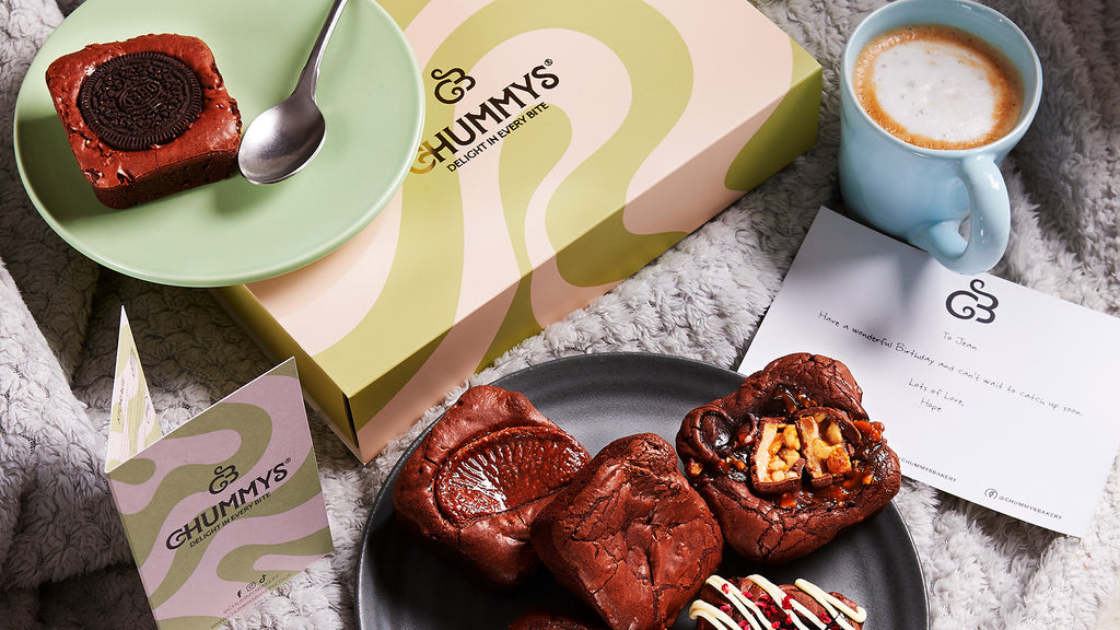 A Chummys box sits on a table. Nearby is a blue coffee mug with coffee and plates filled with brownies