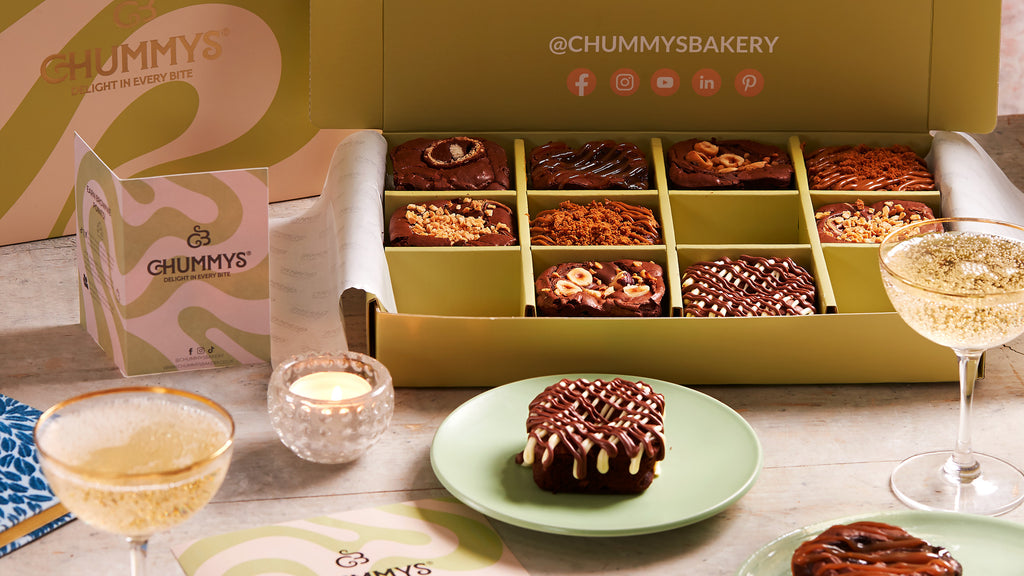 golden box of variety brownies sits on table. Stemmed glasses are placed on the table.