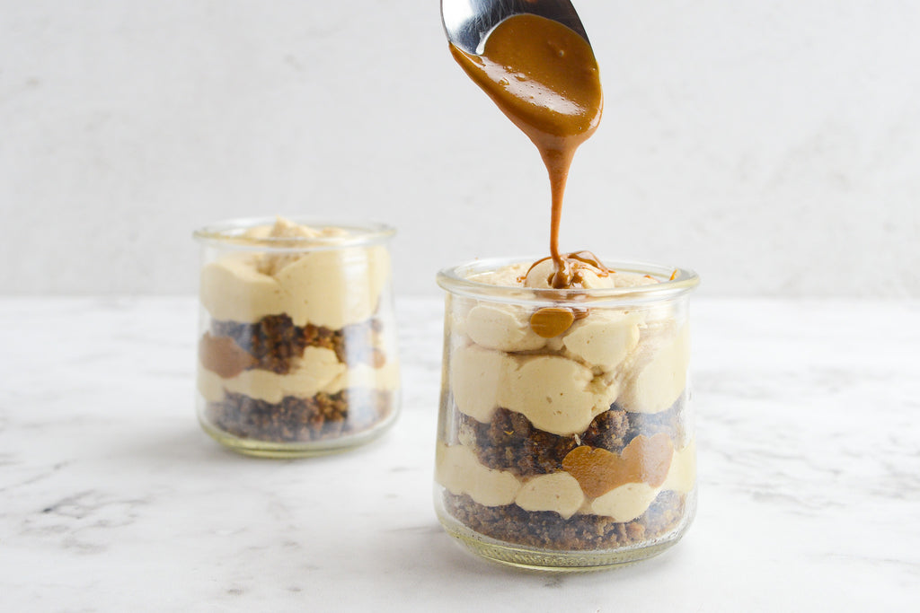 Two cheesecake cups sit on top of marbled table top. A spoon drizzles warm cookie butter on top of one of the cups.