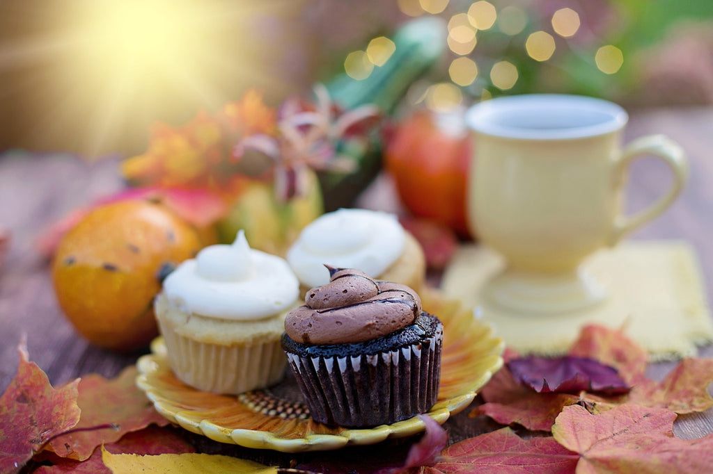 Fall leaf decor covers a small table. On top of the table is a small yellow dessert plate with three cupcakes with a big swirl of frosting. Two are vanilla and one is chocolate. Small fall decor decorates the distant background