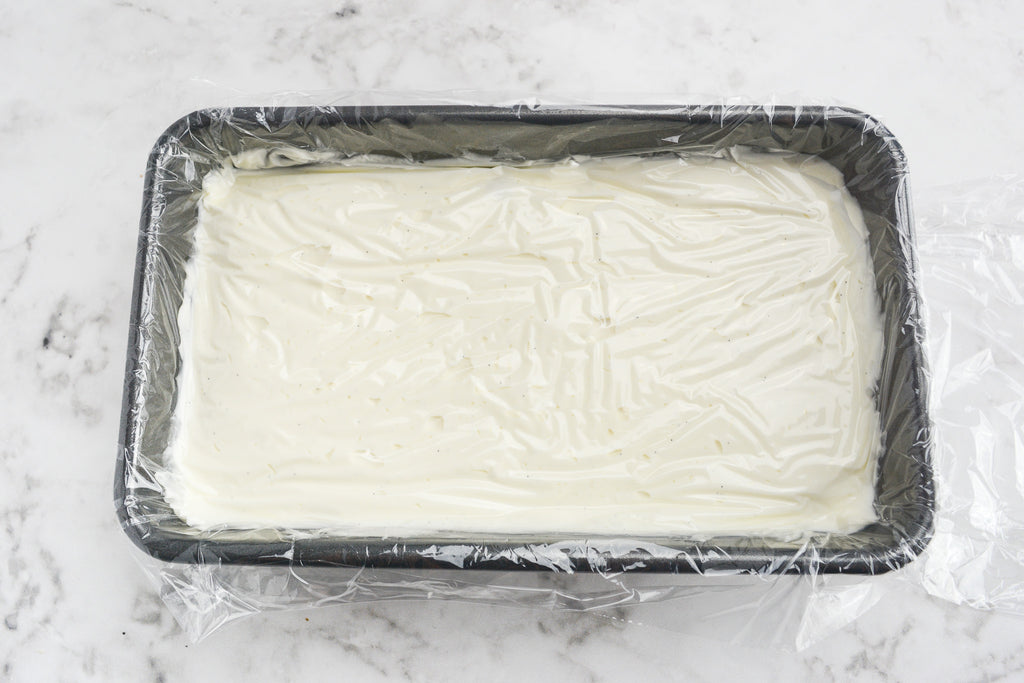 A silver loaf pan is filled with white ice cream. A thin layer of plastic covers the top