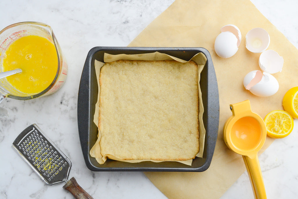 Lemon bar ingredients. In the center a pan prepared with parchment. To the left is a measuring cup with filling and a small grater. To the right is cracked eggs, a lemon juicer and squeezed lemons.